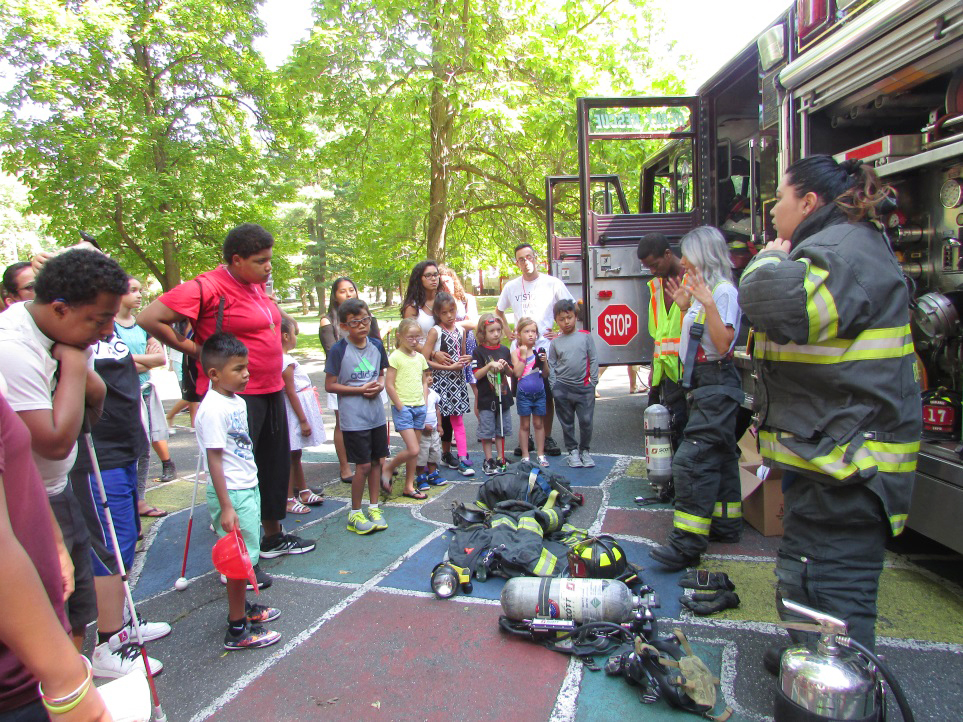firewomen and fireman showing off their equipment next to the fire engine in front of a crowd of children and adults