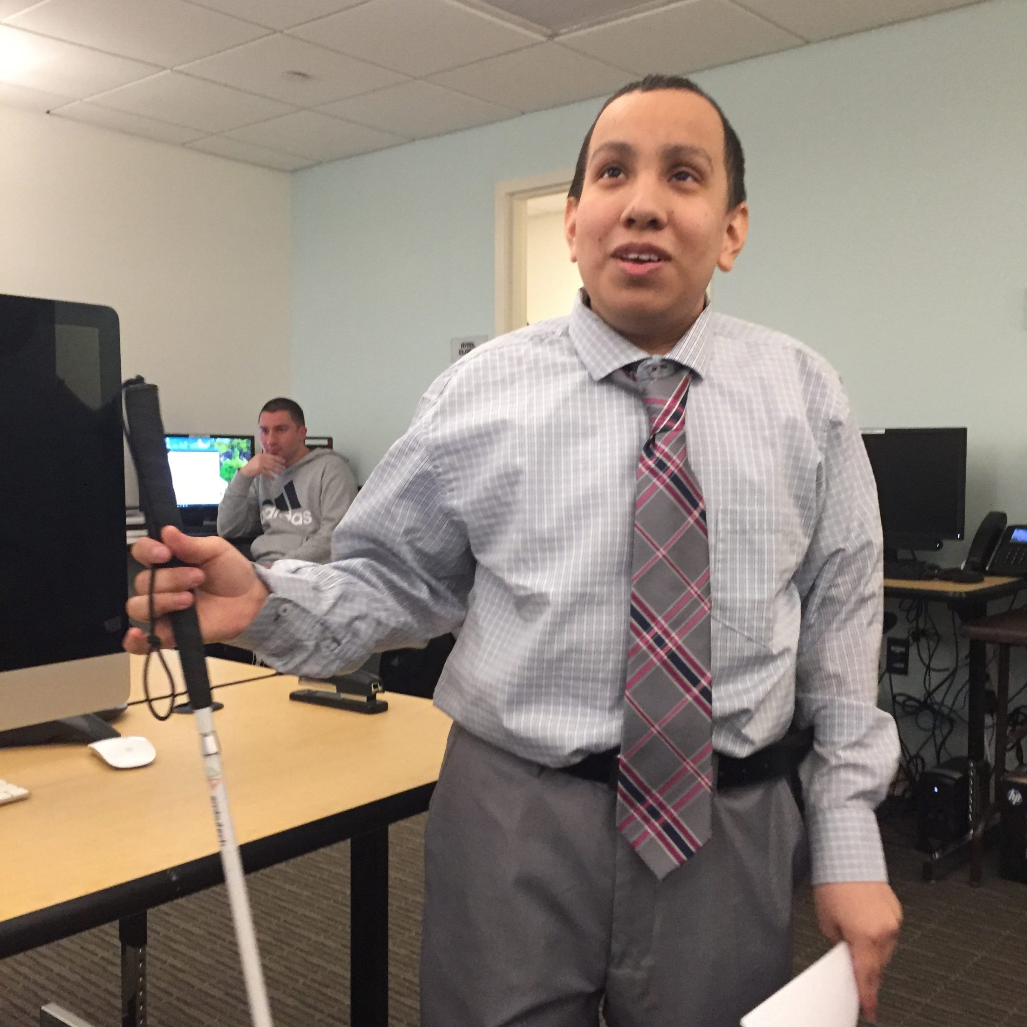 Image description: A young man holding a cane with one hand, and a piece of paper in the other, wearing a light gray button downshirt, gray and red plaid tie, and gray pants stands inside a computer lab.