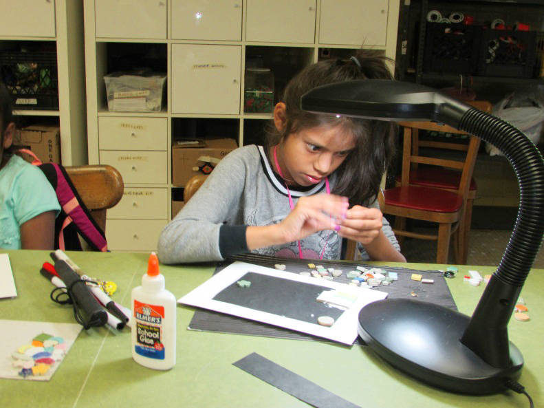 young girl gluing miscellaneous objects to black paper under bright table light