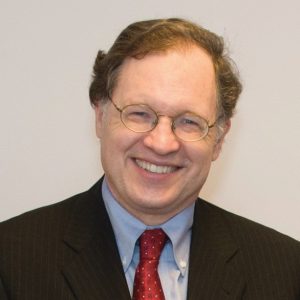 Image description: A man wearing a black suit and red tie stands in front of a white wall and smiles to the camera.