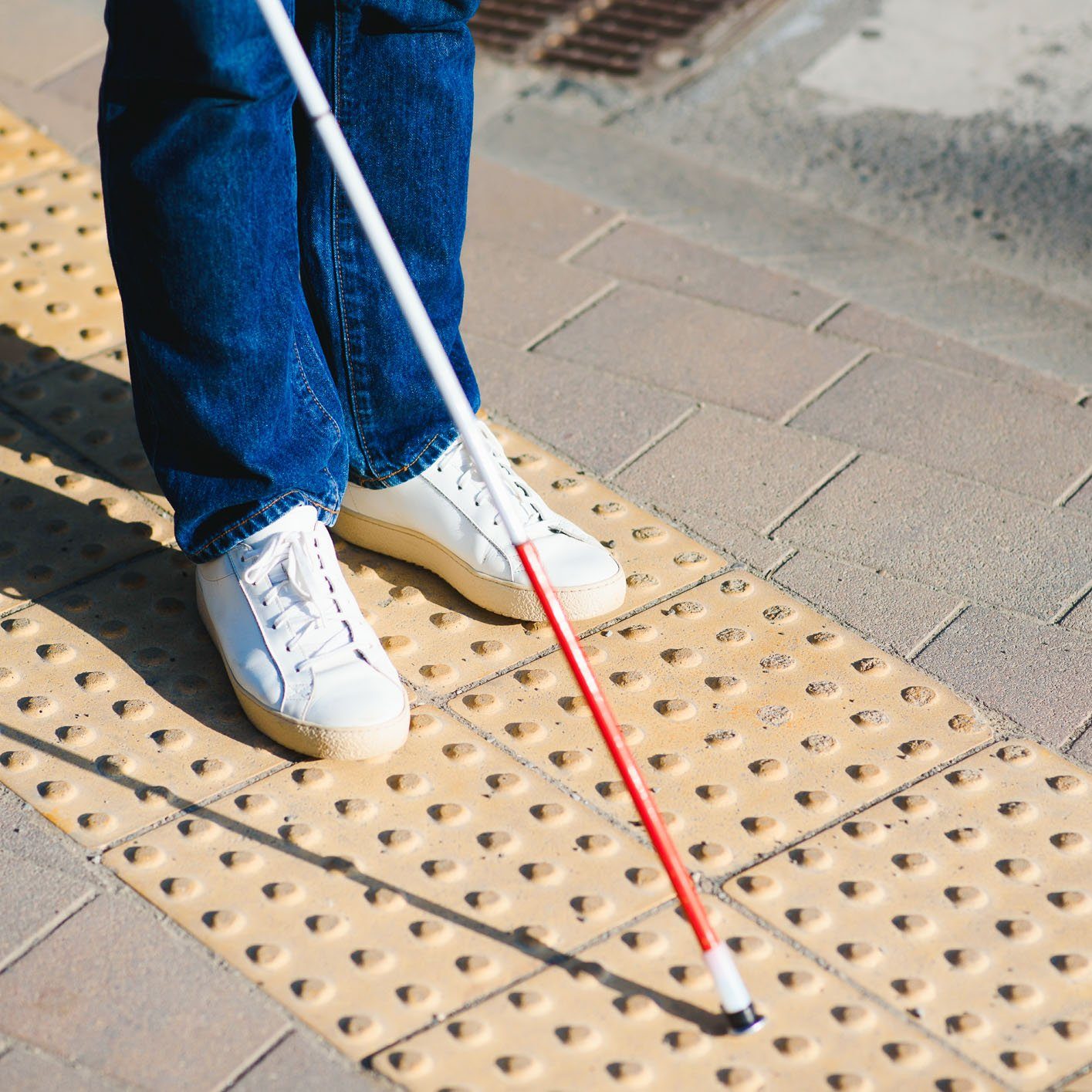 Closeup photo of a person standing on top of yellow truncated domes, as their cane is pointed in front of them.