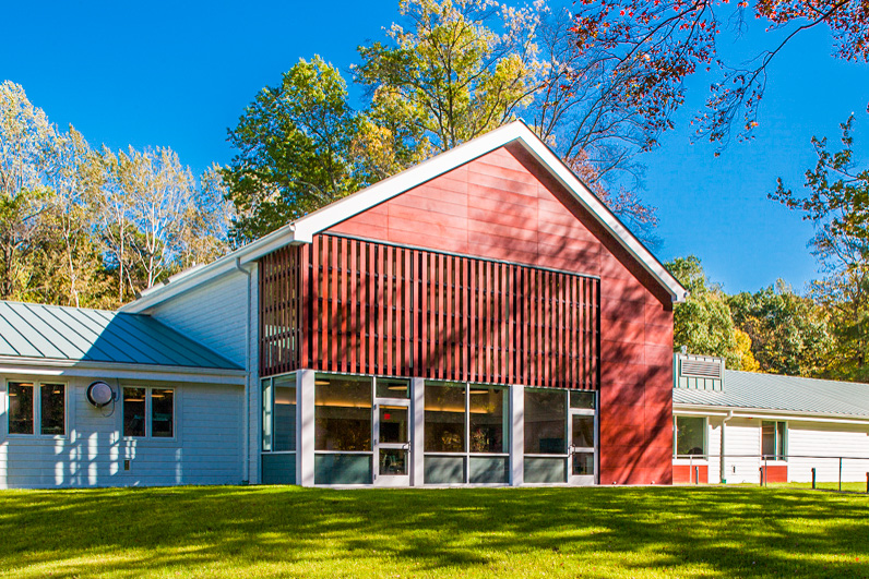 Image description: An exterior view of VISIONS Vocational Rehabilitation Center. A large red barn-shaped building surrounded by trees, on a large grassy field.