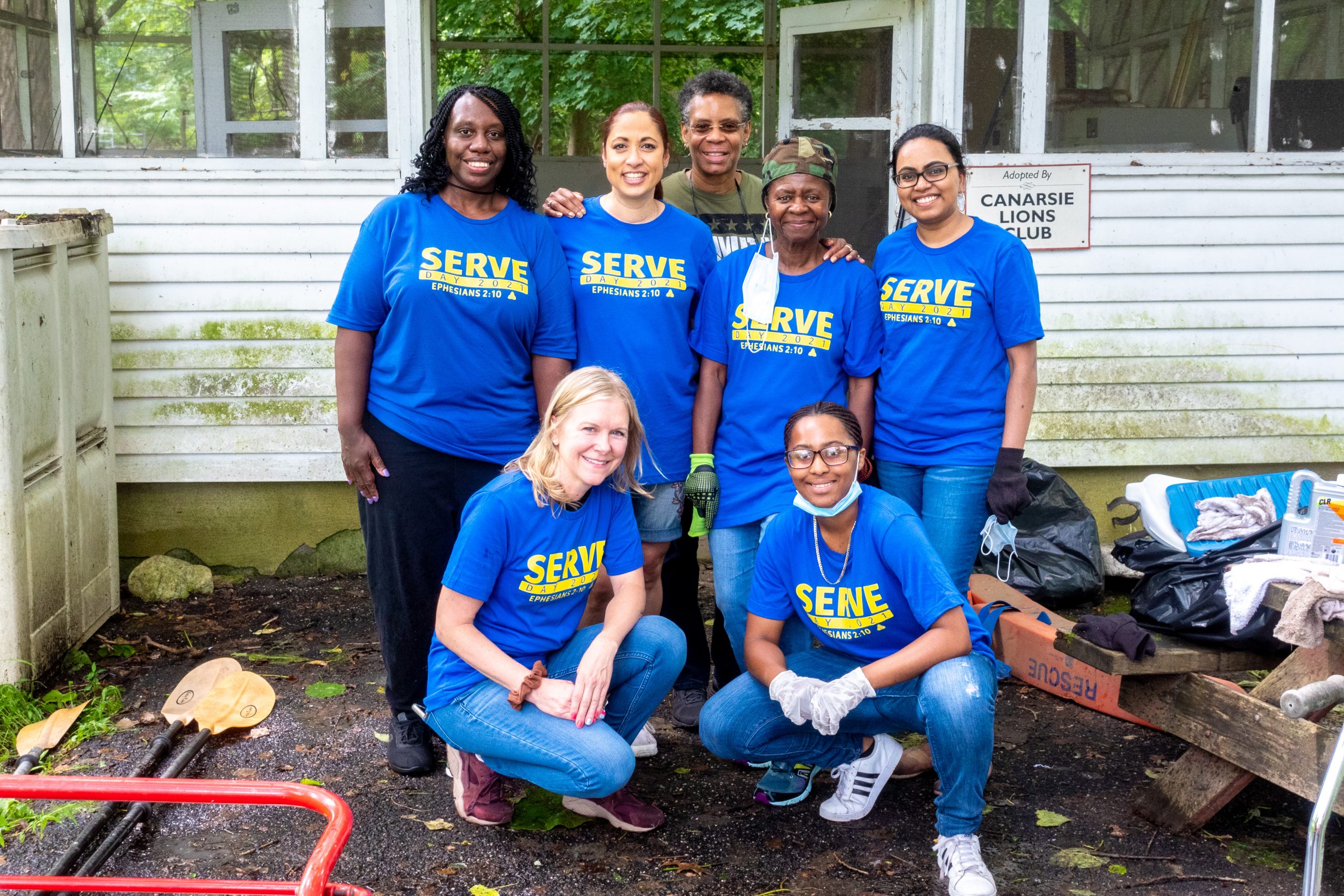 A group of six volunteers wearing blue t shirts stand together, surrounded by cleaning equipment, at VISIONS Center on Aging