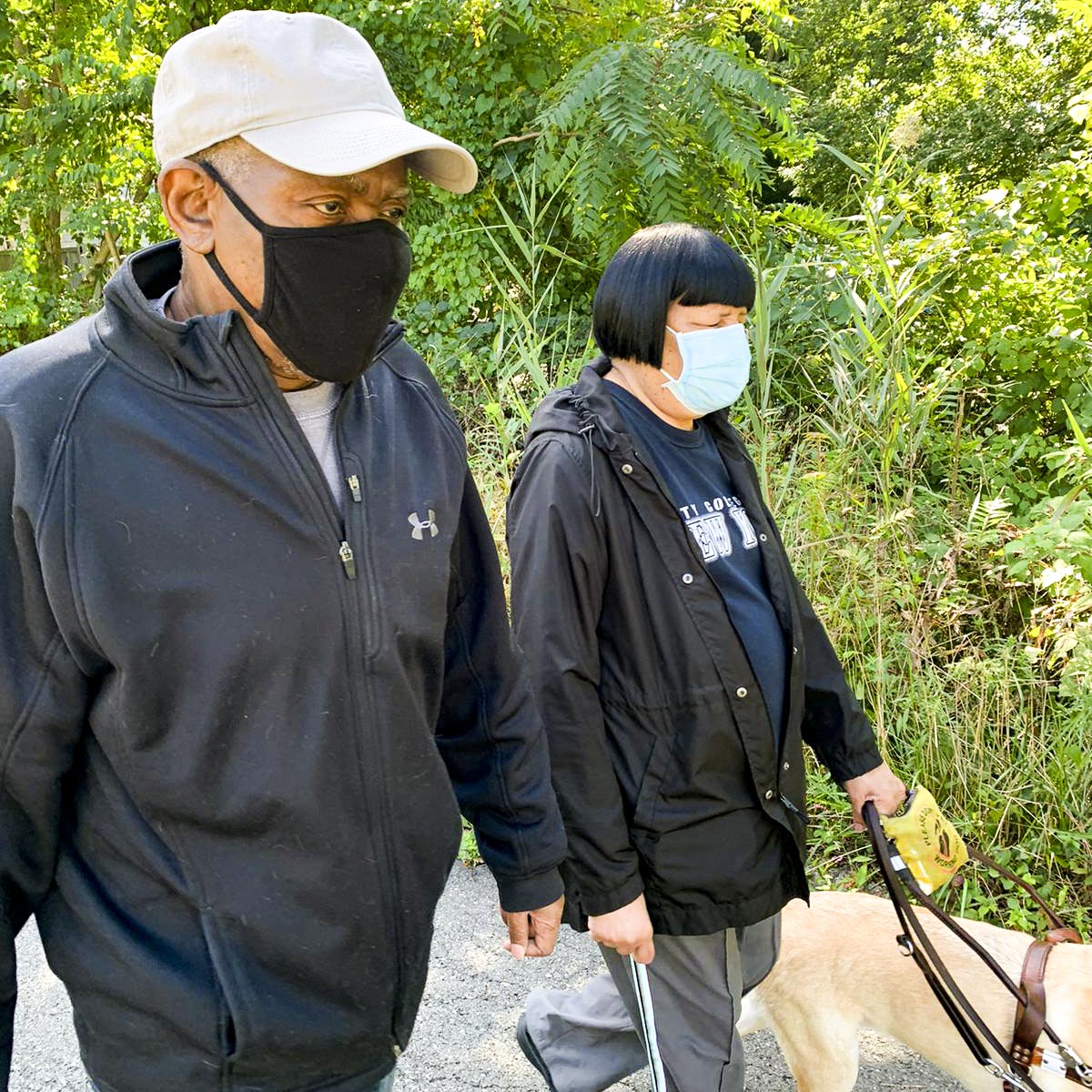 An older participant, with the help of her guide dog, walks along a tree lined street with a caregiver volunteer beside her.