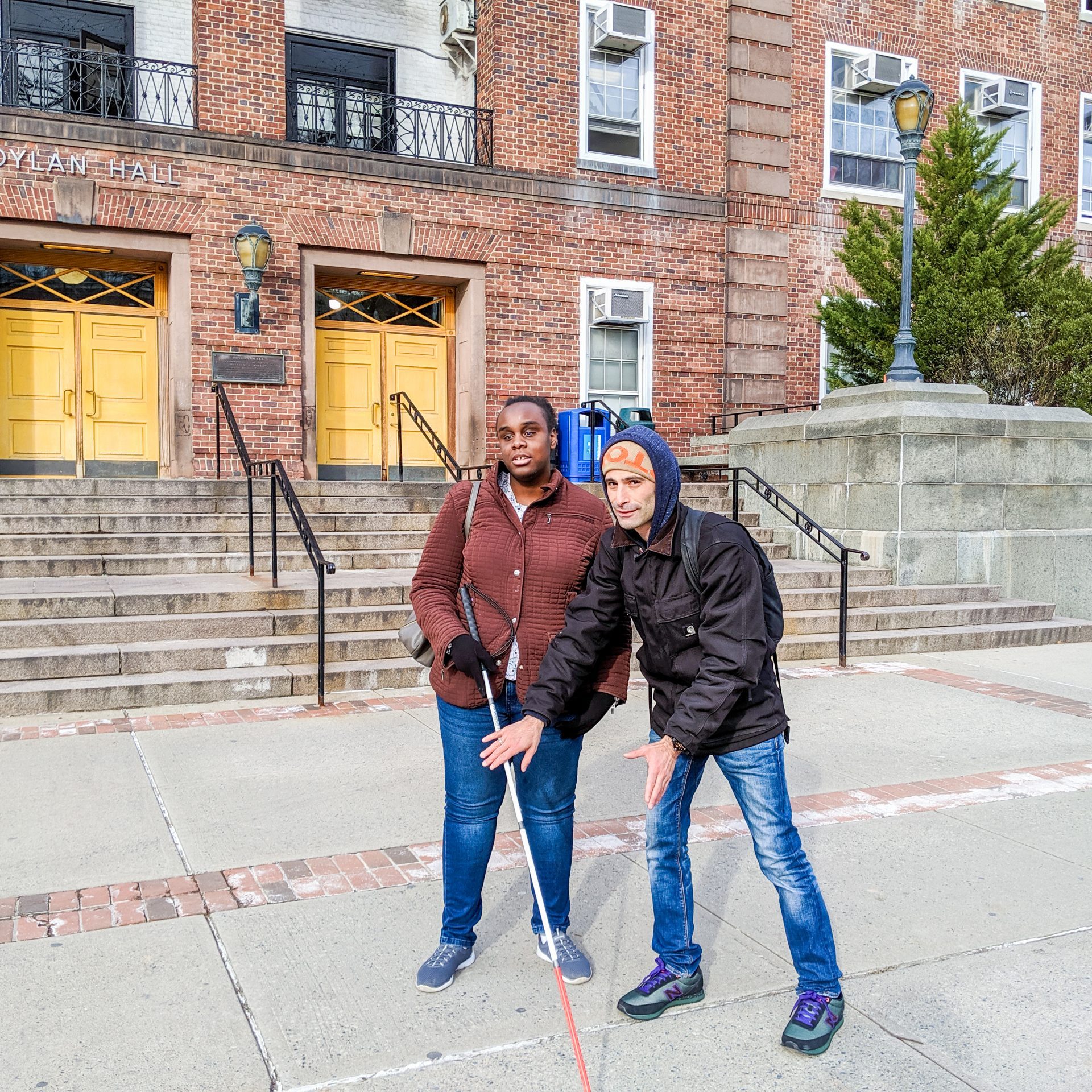 A vision rehabilitation therapist works on proper cane technique with an adult participant outside a large brick building.