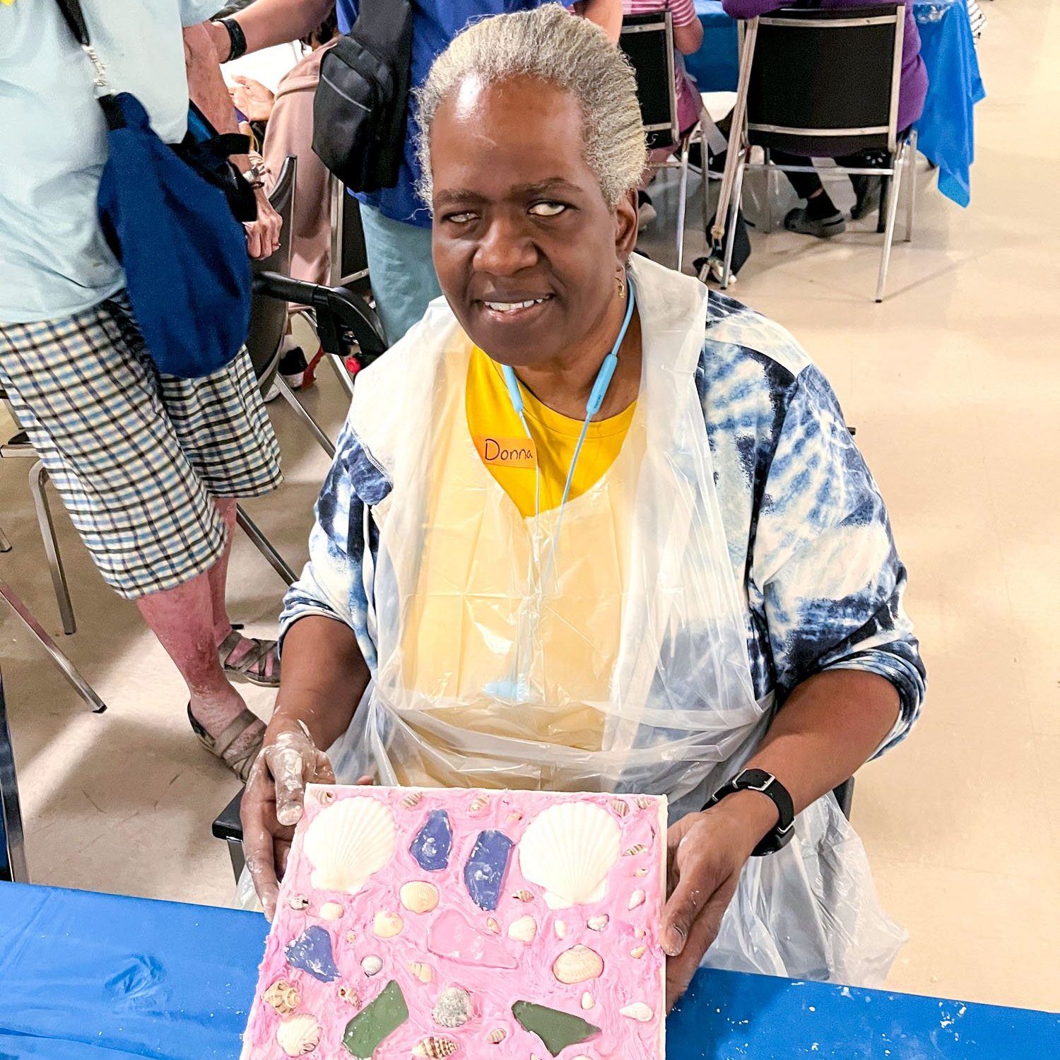 Photo of a smiling older woman sitting at a blue table working on an arts and crafts project.