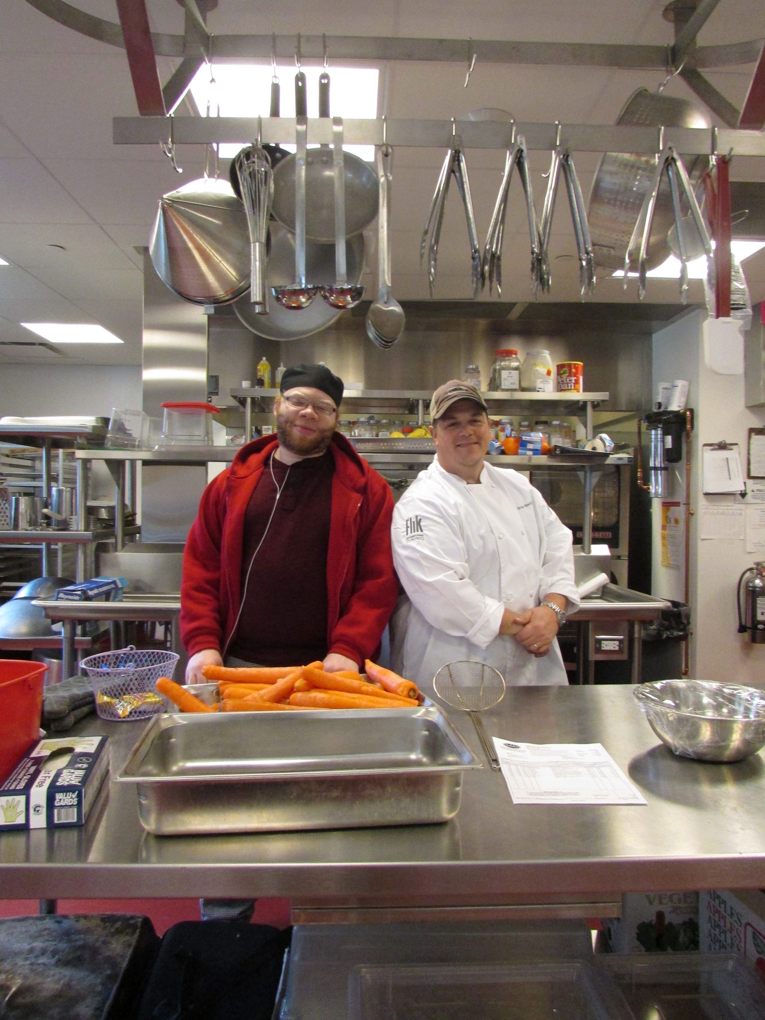 Image description: A man wearing a red sweater and black t-shirt stands next to a man wearing a chef's jacket in a large industrial kitchen. A tray of unchopped carrots sits in front of the men in a metal tray.