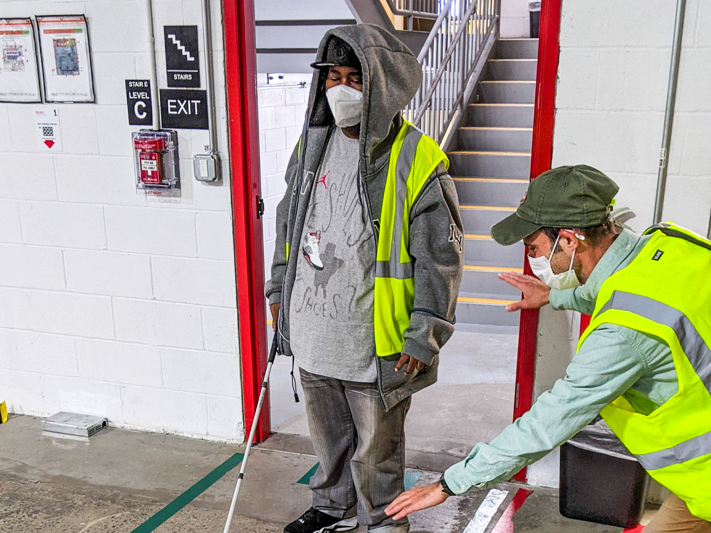 A man wearing a bright yellow safety vest, along with the help of an instructor, learns how to safely navigate a warehouse.