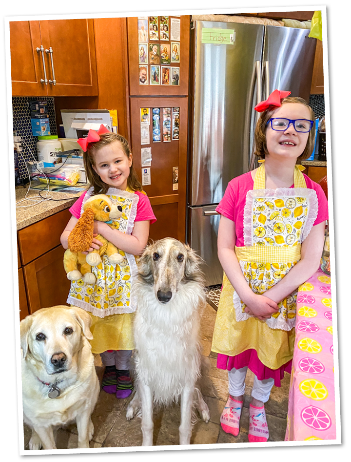 Nuala and Aoife, two young girls wearing pink shirts and lemon aprons stand with their dogs and smile to the camera.